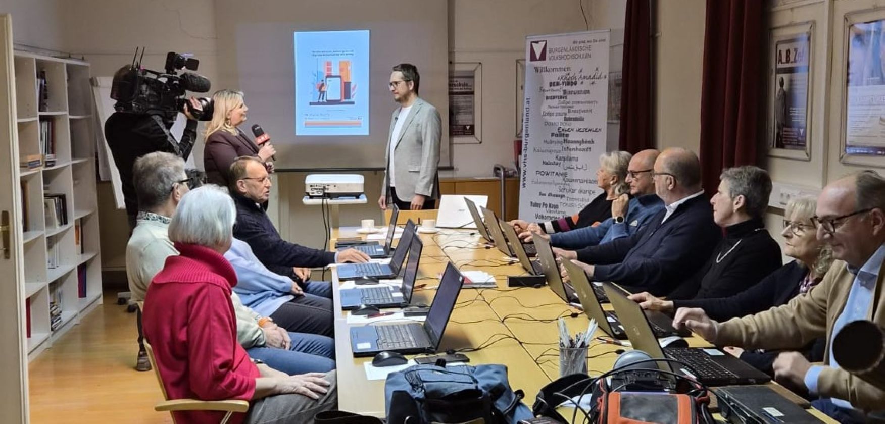 Kursteilnehmer*innen an einem großen Tisch vor Laptops, Kursleiter wird von Reporterin interviewt, Kameramann filmt