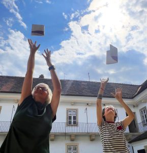 Ursula Foki und Margit Poandl blicken nach oben Richtung Himmel und schnappen sich das neue Kursprogramm, dass ihnen von oben entgegenfliegt.