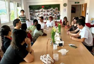 Absolvent*innen des früheren Pflichtschulabschlusses sitzen um einen Tisch. Lehrgangsleitung steht und spricht zu Absolvent*innen. Im Hintergrund sieht man eine Tafel mit "Welcome back"-Schriftzug und vielen Fotos.